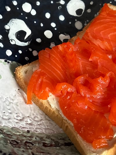 The image displays a close-up view of a slice of bread topped with cream cheese and thinly sliced smoked salmon. The salmon is a vibrant orange-red color, with visible striations indicating its texture. The bread appears to be toasted, with a golden-brown crust and a soft, light-colored interior. The cream cheese is spread evenly beneath the salmon, providing a creamy white contrast.
The toast is placed on a dark surface with a black and white speckled pattern, possibly a plate or placemat. This pattern features irregular white circles and blobs, with some appearing more defined than others. To the right of the toast, a textured, light-colored surface is visible, resembling a rough-hewn material or a patterned fabric. The lighting suggests an indoor setting, with diffused light creating subtle highlights on the salmon and cream cheese. There are no people or animals visible in the image. The focus is entirely on the food item and its immediate surroundings. The overall impression is one of a simple yet appetizing snack or meal. No text is visible in the image.