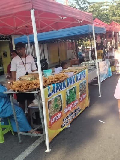 An outdoor street food market stall, identified as "Lapak Pak Kumis," operates under a red canopy tent with white support poles during daylight hours in Situbondo, Indonesia. A prominent yellow banner on the stall advertises "Lumpia," "Biking Cungkil," and "Kue Boler," displaying images of these items, along with contact information: "HUB NO. WA 085338380001 SITUBONDO."

A male adult vendor, with a dark mustache and short beard, wearing a black baseball cap, a white "Nevada" t-shirt, and a lanyard, stands behind the stall, looking towards the left. On the stall's counter, two large circular woven bamboo trays are heavily laden with deep-fried spring rolls (lumpia). Smaller containers with other food items and a light blue plastic container are also present. A young male child is partially visible behind the food display on the right side of the stall.

To the immediate left, a person dressed in light blue long-sleeved clothing and pants is seated on a bright green plastic stool, facing the stall. Their bare feet, with red-painted toenails, rest on the asphalt ground, which has a faded white painted line.

The background reveals a series of additional market stalls with blue and red canopy tents. A black scooter/motorcycle is parked behind a blue tent. Further back, lush green trees are visible, and a building with "ASSAM" marked on its facade is partially seen to the far left. Another adult in a yellow t-shirt is visible under a distant red tent on the right side of the frame.