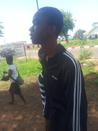 The image shows a street scene in Nsugbe, Nigeria. A young man is in the foreground, wearing a black jacket with white stripes on the sleeves and the word "SPORT" on the chest. He's looking to the left. Behind him, partially obscured, is a second person wearing a striped shirt. To the left of the frame, a woman is carrying a tray of small objects on her head and a bag over her shoulder. She is wearing a white shirt and a dark skirt, with flip-flops. In the background, there's a sign for the "Nwafor Orizu College of Education, Nsugbe - School of Sciences." There's a paved road, green vegetation, and buildings visible in the background. The ground is dirt.