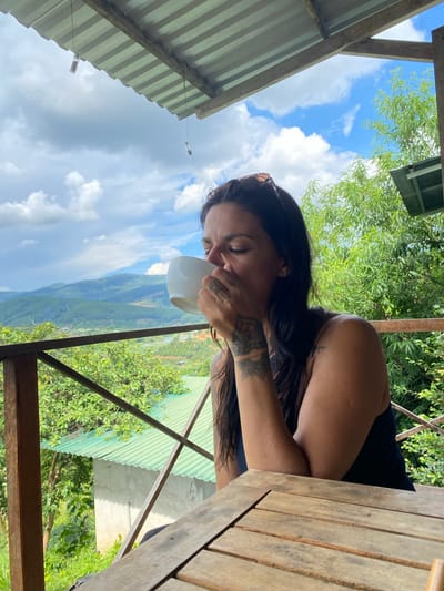 A woman with dark hair and tattoos is drinking from a white mug on a wooden balcony overlooking a lush, green valley with a cloudy blue sky. She is wearing a dark-colored top and has her eyes closed, appearing to enjoy her drink. The setting is rustic, with wooden structures and corrugated metal roofing visible. The lush vegetation and rolling hills suggest a rural or mountainous location. The lighting indicates it is daytime, possibly late morning or afternoon. The overall mood is one of peace and relaxation. The background shows a mountainous landscape, typical of regions like Thôn 3, Vietnam, with visible layers of hills and trees. The architecture of the balcony and the surrounding structures are simple and functional, fitting a natural, perhaps secluded, environment.