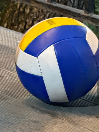 A blue, yellow, and white volleyball rests on a weathered wooden surface, likely a deck or boardwalk. In the background, out of focus, are dark, textured elements that could be trees or fencing. The image captures a close-up view of the volleyball, emphasizing its segmented design and stitching. The lighting suggests it is either dusk or a shaded area. There are no people or actions depicted. The location in the city of Sis, Azerbaijan, is not discernible from the visual information provided in the image.
