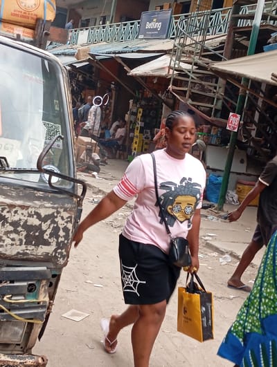 The image depicts a bustling outdoor market or street scene, likely in an urban area during daylight hours.

In the foreground, a woman walks from left to right, her gaze slightly downward. She is wearing a light pink t-shirt featuring a cartoon character with dreadlocks and sunglasses, black shorts adorned with a white spiderweb design on the left leg, and white flip-flops. She carries a black cross-body bag and a yellow and black shopping bag, which has "QUEEN" and possibly "HAIR" visible on it. Her hair is styled in braids.

To the left, a portion of an old, heavily rusted vehicle, possibly a truck or tricycle taxi, is visible, showing its worn headlight and front panel. On top of this vehicle, stacked boxes or crates are visible with text that includes "NATURAL RUBBER BANDS" and "500 G. BANDS".

The background reveals a lively street with multiple people and makeshift stalls or shops. A man in a light-colored, patterned shirt stands near a prominent white ring light on a stand. Several other individuals are scattered throughout the scene, some sitting, some standing, engaged in various activities within the market environment. One man is partially visible on a balcony of a multi-story building further back.

Buildings in the background appear to be multi-storied with metal roofs, exposed structural elements, and balconies with railings. A blue sign with "BetKing" written in white is prominently displayed on one of the buildings. Another red sign with "POS" (likely indicating Point of Sale) is attached to a pole. The ground is unpaved and appears to be dirt or gravel. The overall atmosphere suggests a busy commercial district.