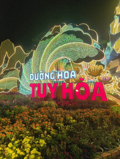 The image depicts a decorative structure and floral display at night, likely in the city of Tuy Hòa, Vietnam, as indicated by the sign reading "Đường Hoa Tuy Hòa," which translates to "Tuy Hòa Flower Street." The structure consists of illuminated, stylized shapes, including curving elements adorned with small, green lights. Above this, decorative gold-colored flowers are visible. A sign with bright red and blue letters reads "Tuy Hoa." A lush flower bed with varying shades of yellow, orange, and red is situated on the right. The background is dark, indicating the night setting.