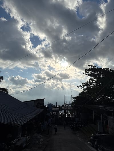 The image shows a street scene in Abang, Indonesia, under a dramatic sky. In the foreground, the left side of the street is lined with buildings featuring corrugated metal roofs, appearing to be market stalls or small shops. A person is walking down the paved street in the center, away from the viewer. The right side of the street is dominated by a dense tree, silhouetted against the sky, and more low-lying structures with covered roofs. Further down the street, a swing set is visible, and beyond that, structures with domes or pointed roofs suggest a religious building, possibly a mosque. The sky is filled with a mix of dark, heavy clouds and patches of bright blue sky, with sunlight breaking through in rays. Overhead power lines are strung across the scene.