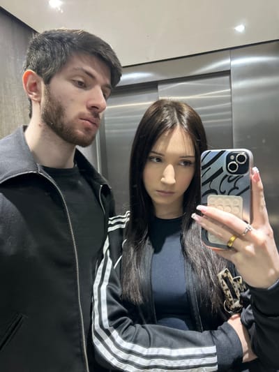 This image captures a mirror selfie of two individuals, a male and a female, taken inside an elevator located in Cachan, France. The male, positioned on the left of the frame, has short brown hair and a beard. He wears a dark, zippered jacket over a dark top and is looking downwards and to the right. The female, positioned to the right and slightly behind the male, has long, dark brown hair. She wears a black Adidas track jacket with white stripes on the sleeves over a dark, form-fitting top. Her right hand, adorned with a gold-colored ring on the ring finger and a silver-colored ring on the middle finger, holds a silver-colored iPhone equipped with a clear case featuring a blue abstract pattern and an integrated pop-it fidget toy. A dark and light patterned accessory is visible near her left wrist. Both subjects exhibit neutral or slightly somber expressions, with their gaze directed away from the smartphone camera. The environment is characterized by brushed metallic elevator walls and overhead lighting.