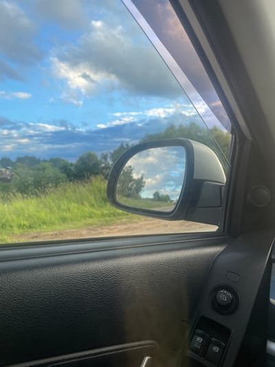 The image is a close-up view from inside a car, looking out the side window and into the side mirror. The scene outside is rural, with green fields and trees under a cloudy blue sky. The side mirror reflects the sky and a line of trees.

Inside the car, the focus is on the door panel. We see the textured black plastic of the door, the window frame, and the edge of the car's interior trim. A side mirror, mounted on the exterior of the car, is visible through the window, and its reflection is captured in the mirror itself. On the door panel, there are control buttons, including a circular dial with directional arrows and two rectangular buttons, one labeled "AUTO DOWN."

The lighting suggests it is daytime, possibly late afternoon, given the soft light and the presence of clouds. The overall impression is one of a quiet moment inside a vehicle, observing the surrounding countryside. There is no discernible text in the image, other than the labels on the window control buttons. The setting appears to be rural Russia, based on the provided location context.