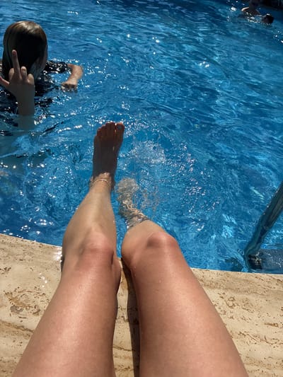 In this high-angle shot, two pairs of legs are visible, one pair belonging to a person seated on the edge of a swimming pool, and another pair of legs, belonging to a child, are submerged in the bright blue water. The seated person's legs are light-skinned, with one foot partially in the water, showing a toenail painted a pale blue. A thin, gold anklet is visible on this foot. The child, with dark hair, is partially visible in the pool, with their head and arms above the water. Their left hand is raised, and their fingers are extended in what appears to be a peace sign. The pool water is clear and reflects the sunlight, creating a shimmering effect. The edge of the pool is a textured, beige material, possibly travertine tile. The scene suggests a warm, sunny day, likely in the afternoon given the bright sunlight and the shadows cast on the water. The overall mood is relaxed and summery, indicative of a vacation or leisure activity. There is no visible text in the image. The location context is Manavgat, Turkey.