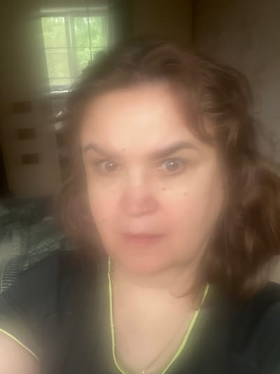 A medium shot captures a woman from the chest up, facing forward. She has light skin, dark brown wavy hair, and is looking directly at the camera with an intense expression. Her eyebrows are dark and arched. She is wearing a dark top with a neon yellow trim. A delicate silver necklace is visible. The background is softly lit and out of focus, suggesting an indoor setting. To the left, a window with light green curtains reveals blurry greenery outside, hinting at trees and foliage. The overall impression is that of a dimly lit room with natural light filtering in from the window. There is no identifiable landmark or specific object in the scene that would suggest the location of Tula, Russia.