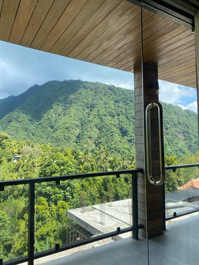 The image depicts a view of a lush green mountainside from a balcony in Sawan, Indonesia. The foreground features a modern balcony with a glass railing, a wooden ceiling, and a prominent silver door handle. To the right, a wooden-paneled column supports the structure, and a light fixture is visible. Below the balcony, construction is underway on a concrete structure, with rebar protruding from the surface. The middle ground is dominated by a dense canopy of tropical trees, including palm trees, interspersed with the green slopes of the mountains. In the distance, the imposing green mountains rise, their surfaces covered in foliage. The sky above is partly cloudy, with patches of blue visible. The scene conveys a sense of natural beauty and ongoing development in a tropical setting.