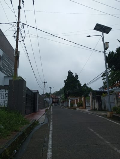 A wet, uphill street leads through a residential area under a cloudy sky. Power lines crisscross above the narrow road, connecting poles along the street and a solar-powered streetlight. Lush green vegetation sprouts along the roadside. Homes with fences and gates line the street, some old and some recently constructed. Tall trees are visible in the background.