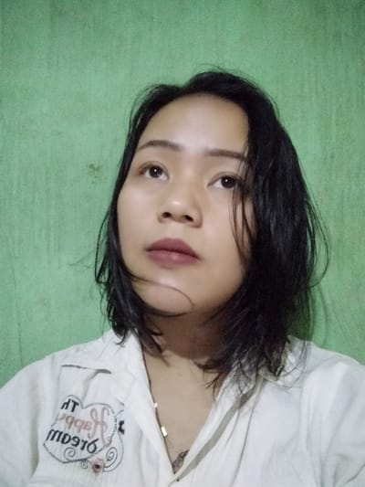 A young Asian woman with short, dark, slightly wet hair looks upward. She has light brown skin, dark eyes, and full lips coated with a mauve-toned lipstick. She wears a white collared shirt with a decorative heart and text graphic on the left side. A simple necklace is visible beneath the collar. The background is a solid, slightly textured green wall. The lighting is soft, giving the image a slightly muted quality.