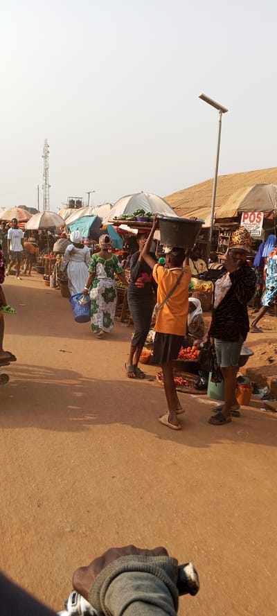 This image captures a lively outdoor market scene in Ketu Adieowe, Nigeria, under a bright, clear sky. The ground is a reddish-brown dirt path, suggesting an unpaved road or walkway.

Numerous people, mostly women, are visible, engaged in daily market activities. In the mid-ground, a person wearing an orange shirt and black shorts balances a large, dark bucket on their head, walking through the crowd. Another woman, dressed in a patterned white and green attire, carries a blue plastic basket. Market stalls line both sides of the path, shaded by various canopies and umbrellas, beneath which produce like fruits and vegetables are displayed. One stall clearly shows a "POS" sign, indicating a point-of-sale service is available.

In the distance, a tall telecommunications tower is visible against the light sky. Closer to the right, a modern solar-powered street light stands. The light suggests it's daytime, possibly morning or afternoon, with noticeable shadows. The immediate foreground features a dark-skinned hand, likely the photographer's, gripping a bicycle handlebar, implying the photo was taken while riding or stationary with a bike. The overall atmosphere is one of a busy, active local marketplace.