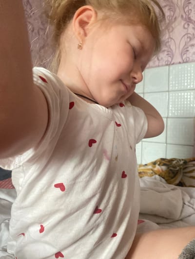 A young child with blonde hair is sleeping on their side, facing left. They are wearing a white t-shirt adorned with small red hearts. The child's eyes are closed, and their face appears relaxed. A delicate gold earring is visible on their left ear. The background shows a tiled surface and a patterned fabric, possibly a bedspread or blanket. The scene suggests a moment of rest or sleep, possibly during the daytime given the lighting. The overall mood is peaceful.