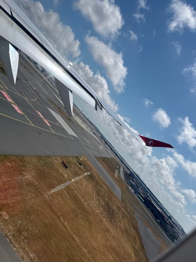 The image, taken from an airplane window, offers a perspective of an airport and its surroundings in Arnavutköy, Turkey. The foreground is dominated by the airplane's wing, gleaming in the sunlight, with its distinctive Turkish Airlines logo visible on the wingtip. Below the wing, the airport tarmac stretches out, marked with various lines and signals, suggesting active runways and taxiways. Beyond the tarmac, dry, brown fields with some scattered vegetation are visible, indicating a semi-arid or seasonal landscape. In the distance, a sprawling urban landscape with numerous buildings and structures can be seen, likely part of the airport infrastructure or the city itself. The sky is a vibrant blue, dotted with fluffy white clouds, suggesting clear weather conditions. The angle of the photograph suggests the plane is either taking off or landing.