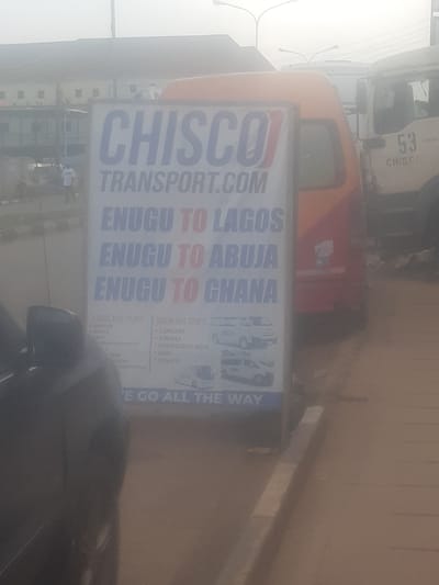 The image shows a sign for "CHISCO TRANSPORT.COM" which states routes such as "ENUGU TO LAGOS", "ENUGU TO ABUJA", and "ENUGU TO GHANA". The sign also lists bus stops for Lagos and Abuja. In the background, there are several vehicles, including what appear to be buses and trucks, suggesting this is a transportation hub or terminal. The environment appears to be an urban setting, possibly a busy street or a depot, with overcast weather conditions indicated by the hazy sky. The sign is positioned outdoors, on a sidewalk or roadside, and a portion of a black vehicle is visible in the foreground on the left. The sign is slightly tilted, and some parts are obscured by glare. The overall impression is that of a functional advertisement for a transport company operating within and beyond Enugu, Nigeria.