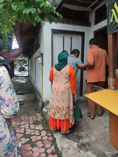 An outdoor scene in Lamongan, Indonesia, captures several people standing along a narrow pathway between buildings, suggesting an alleyway or a residential street. The ground consists of concrete, with some areas appearing damp, and reddish, patterned paving stones are visible on the right. Green foliage peeks in from the left side of the frame.
In the mid-ground, a person wearing a dark green hijab, a light-colored top with a floral pattern, and a vibrant orange ruffled skirt stands with their back to the viewer. They carry a dark green handbag. Beside them, a man in a grey t-shirt also faces away. Further into the scene, a man in an orange long-sleeved shirt and brown pants stands near a white-framed doorway or window, seemingly entering or waiting. Another person with dark hair is visible between the grey-shirted man and the doorway. In the foreground, only the legs of another individual, clad in patterned pants, are partially visible.
A dark green banner with a yellow, stylized letter or logo is partially visible at the top left. Architectural elements include white walls, dark wooden frames, and some brown lattice work. In the distance down the path, a motorcycle can be seen. The setting appears to be daytime, likely overcast given the soft lighting. The people seem to be gathered or waiting, with their attention directed forward and slightly to the right.