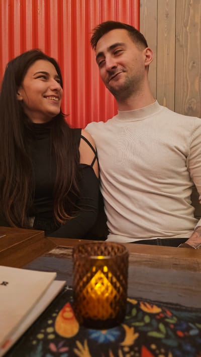 The scene, located indoors in Aschaffenburg, Germany, depicts two adults seated at a wooden table. On the left, a female subject with long dark hair, wearing a black long-sleeved top featuring a criss-cross cut-out design on the left shoulder, is smiling and gazing towards the male subject. On the right, a male subject with short dark hair and light facial stubble, wearing a light cream, high-neck sweater, exhibits a faint smile while looking downwards and to his right; his left arm is positioned behind the female subject. His right forearm displays visible tattooing. In the foreground, centered on the table, is a dark amber, diamond-patterned glass candle holder emitting a warm orange light from an internal source, resting on a dark mat adorned with a colorful, abstract floral pattern. A white booklet with a partially visible 'R' on its cover is also present on the table's left side. The background consists of a vertical corrugated red wall behind the female subject, transitioning to a vertical wooden plank wall behind the male subject.