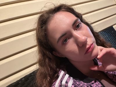 The image is a selfie of a young woman. She has long, wavy brown hair and is looking directly at the camera. Her skin appears smooth and slightly flushed, with natural makeup. She is wearing a black jacket with pink and white geometric patterns and the iconic Adidas stripes on the sleeves. Her right hand is touching her chin, and her expression is neutral.
The background consists of horizontal, cream-colored panels, suggesting an indoor or covered outdoor setting with textured walls. To the right, a black woven material is visible, possibly part of a chair or furniture. The lighting appears to be warm and possibly artificial, with some shadows cast on her face and the background.
While the image itself doesn't provide explicit clues about the time of day or weather, the lighting suggests it could be late afternoon or evening, or an indoor setting with warm lighting. The woman's relaxed posture and neutral expression don't indicate any specific event or strong emotion. The location context provided is Zavodoukovsk, Russia. There is no visible text in the image.