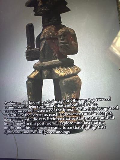 The image displays a carved wooden statue, identified by accompanying text as "Arobinagu," "Ahobinagu," or "Obinagu," a revered cosmic force in Igbo spirituality, culturally associated with Nnobi, Nigeria. The statue depicts a dark-toned male figure, likely seated or squatting, with bent legs. He wears a distinct, possibly tiered, headpiece. His left hand firmly grasps a short, cylindrical object. His right hand is raised, with fingers partially discernible. A prominent, textured band encircles his waist. Traces of red and golden pigment are visible on the figure's chest, waist, and legs. The statue and its descriptive text are presented against a plain light grey background, suggesting a museum display, gallery setting, or digital presentation. The image appears to be a photograph of a screen or print, exhibiting minor reflections and a soft focus.