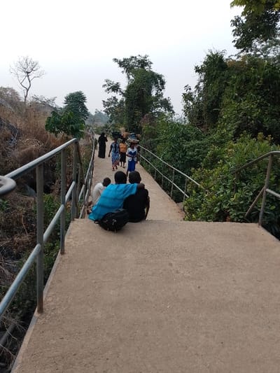 The image captures an outdoor scene featuring a concrete pathway or bridge with metal railings, surrounded by natural vegetation. In the foreground, two individuals are seated on the path with their backs to the viewer. One person is wearing a bright blue garment over a dark top, while the other wears a dark top. A black backpack rests on the path next to them. Further along the pathway, several people are walking away from the camera. Among them, one person is dressed in a long dark robe. Other individuals, primarily women, are visible, some carrying baskets or bundles on their heads, a practice known as head-panning. Their attire includes various colored clothes, such as blue, orange, and white.
The path is flanked by dense green foliage, bushes, and trees on the right side. On the left, the terrain appears drier, with sparser, brownish vegetation, possibly rocky outcrops, and a distinctive bare tree with many branches silhouetted against a light, hazy sky. The overall lighting suggests either an overcast day or an early morning/late afternoon time, as there are no strong shadows. The scene depicts a moment of daily activity, with people traversing the path, while others take a moment to rest.