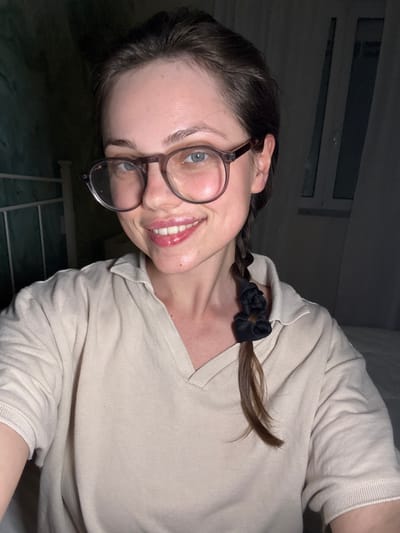 A young adult female, identified as being in Scafati, Italy, is depicted in a dimly lit indoor setting. She has light skin, medium-brown hair pulled back, with a single braid secured by two black scrunchies draped over her right shoulder. She wears clear-framed, round eyeglasses and a light beige or cream-colored short-sleeved V-neck knit top. Her expression is a wide smile, displaying her teeth and a glossy appearance on her lips. Her light-colored eyes are visible through her glasses, reflecting light. The background includes a white metal bed frame headboard against a dark, possibly green wall on the left, and a dark window with visible panes partially obscured by a light curtain on the right. A white surface, likely a bed, is visible in the lower right.