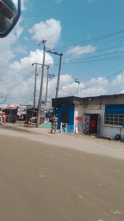 The image depicts a street scene in Effurun, Nigeria, with a sandy road leading to a row of low buildings and commercial establishments. Overhead power lines crisscross the sky, which is partly cloudy. A man is walking along the side of the road, near a blue and white storefront advertising phone accessories. To the left, there are open-air stalls with goods displayed, and people are present in that area. The buildings have a somewhat weathered appearance, and the scene has a general atmosphere of a typical urban street in a developing country.