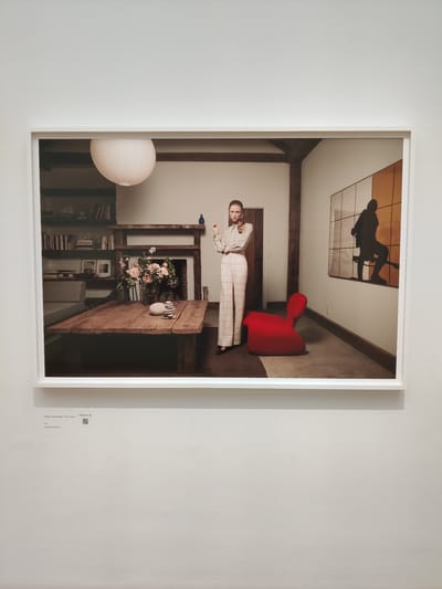 This is a photograph of a framed artwork displayed on a white wall. The artwork itself depicts an interior scene with a woman standing in the center. She is wearing a patterned blouse and high-waisted checkered pants. In the background, there is a rustic wooden table with a floral arrangement in a vase, a fireplace, bookshelves, and a framed abstract artwork on the wall. A bright red chair is placed on the floor to the right of the woman. Below the framed artwork, a label with text and a QR code is visible. The location is the city of The Hague, Netherlands.