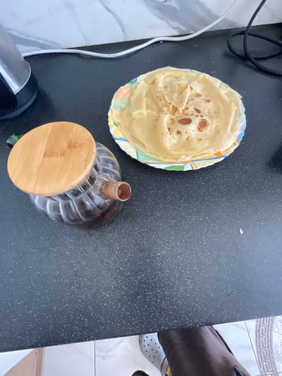 A high-angle shot captures a breakfast scene on a speckled black countertop in Bol'shoe Zaborod'e, Russia. In the foreground, a clear glass carafe filled with a dark liquid, possibly coffee or tea, is topped with a round, light-brown wooden lid. To the right of the carafe, a stack of thin, golden-brown crepes rests on a plate with a colorful rim featuring floral patterns. The background shows a white, marble-patterned backsplash and coiled black and white electrical cords. The scene is illuminated by overhead lighting, casting subtle shadows.