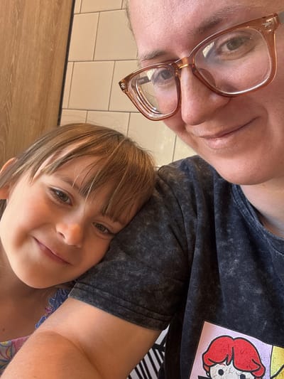 A medium shot captures a young girl resting her head on the shoulder of an adult who is wearing glasses. The girl is on the left, with her eyes open and a smile on her face. Her hair is brown and falls across her forehead. She is wearing a pink and blue floral dress. The adult is on the right, their face partially visible, wearing large, brown-rimmed glasses. They are wearing a dark gray t-shirt with a graphic print of cartoon characters. The background shows a tiled wall and a wooden surface. The image appears to be a selfie taken indoors.