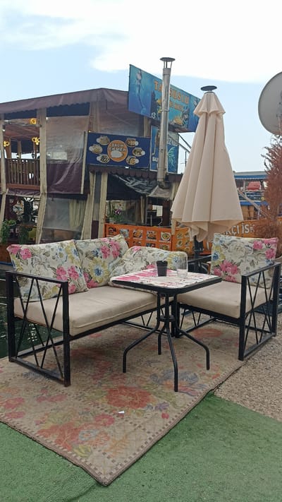 A waterside cafe with outdoor seating is depicted. A two-seater bench with floral cushions and a matching armchair are arranged around a small, square table. On the table, there is a dark cup and a clear glass. A large, cream-colored umbrella is partially open behind the seating. A floral rug is spread on the ground beneath the furniture. In the background, a structure that appears to be a boat or a pier-side building houses a cafe with windows and signage. One sign features a mermaid and Turkish text. The cafe is situated by the water, with the water visible in the foreground. The overall atmosphere suggests a relaxed, outdoor dining experience.