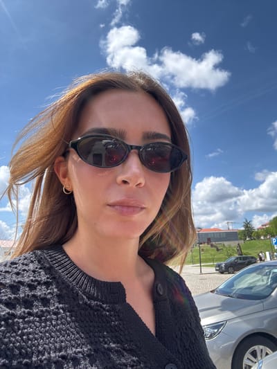 A low-angle, slightly blurred, color photograph shows a woman wearing sunglasses and a dark, crocheted sweater. She is outdoors on a sunny day, likely in Mogadouro, Portugal. Above and behind her is a bright blue sky dotted with white clouds. Reflections visible in her sunglasses indicate the presence of nearby buildings. The side of a silver car is partially visible on the right. A street, green grass, a parking sign, a palm tree, and buildings can be seen in the background, further suggesting an urban setting in Mogadouro.