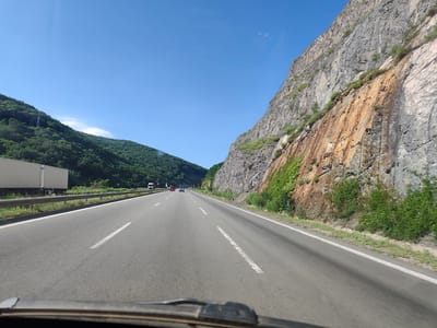 The image shows a highway with a sheer rock face on the right and a densely vegetated hillside on the left. The highway is paved with asphalt and has white lane markings. There are several vehicles on the road, including cars and trucks. A large white semi-trailer truck is visible on the left side of the road. The sky is clear and blue. The rock face on the right is textured and shows evidence of erosion, with some reddish-brown staining. Green vegetation is growing on the rock face and along the sides of the road. The scene appears to be a scenic route, possibly in a mountainous or hilly region. The presence of a highway suggests a location outside of a dense urban area, and the natural landscape indicates a rural or semi-rural setting. Given the prompt's mention of Malo Buchino, Bulgaria, this highway is likely a route passing through or near the village, potentially connecting it to other areas.