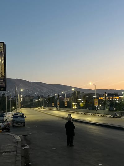The image captures a twilight street scene in what is identified as Al-Qanawat Municipality, Syria. The sky transitions from a soft blue-grey on the left to a warm, pale orange and yellow hue towards the right horizon, indicating either sunset or sunrise.

In the mid-ground, a prominent, dark mountain range stretches horizontally across the frame, with numerous small lights scattered on its slopes, suggesting residential areas built into the mountainside.

The foreground is dominated by a wide, paved road illuminated by a series of streetlights that cast warm glows. On the right side of the road, a lone figure, appearing to be a man dressed in dark clothing and a light-colored hat, walks away from the camera. Further down the road on the right, a light-colored truck is visible.

Along the edges of the road, multiple streetlights are active, and various buildings with illuminated windows can be seen. On the bottom left, a section of a billboard is visible, with faint, partially obscured text including what appears to be "3551" and "pb". The overall impression is that of an urban or semi-urban environment nestled against natural topography, with artificial lights beginning to dominate as daylight fades.