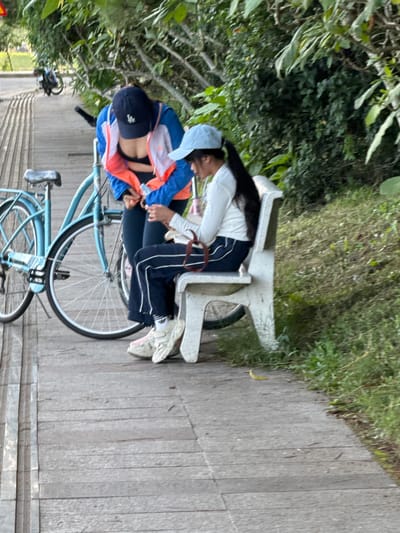 The image captures a scene on a wooden walkway in Xuan Huong Lake area, Da Lat, Vietnam. Two individuals are present. One person, wearing a blue and orange jacket and a dark baseball cap, is leaning over a light blue bicycle. This individual appears to be interacting with a smartphone or a similar device. The second person, seated on a white concrete bench, is wearing a white long-sleeved shirt, dark pants with white stripes, and a light blue baseball cap. This individual is also looking down at a device, possibly a phone, and holding a pink cup with a straw. The bicycle is parked next to the walkway. The environment is characterized by lush green foliage and trees in the background, suggesting a park or natural setting. The lighting indicates it is daytime.