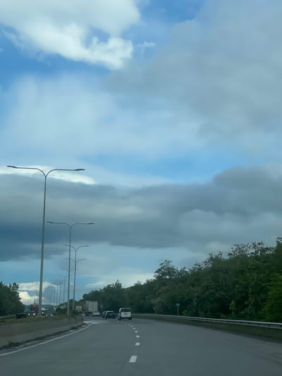 The image depicts a roadway in Mrgvali Chala, Georgia. The sky is filled with a mix of blue and gray clouds, with some appearing to be storm clouds. Along the roadside, there are evenly spaced light poles. A row of trees borders the right side of the road. There are a few vehicles on the road, including what appears to be a truck and several cars.