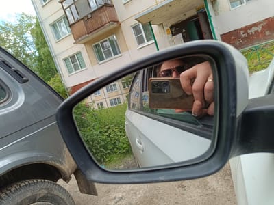 A rear-view mirror reflects a person holding a smartphone, taking a selfie. The person is wearing sunglasses and a ring on their left hand. The background reflected in the mirror shows a residential building with multiple windows and balconies, and lush green bushes with some pink flowers. To the left of the mirror, the side of a grey car is visible, with its tire and mudguard showing. To the right of the mirror, the white body of another car is visible. The setting appears to be outdoors, likely in a residential area, possibly in Arkhangelsk, Russia, given the architectural style of the building. The lighting suggests it is daytime, and the weather appears to be overcast, as there are no strong shadows. The overall scene is casual and everyday, with the focus on the person capturing a moment.