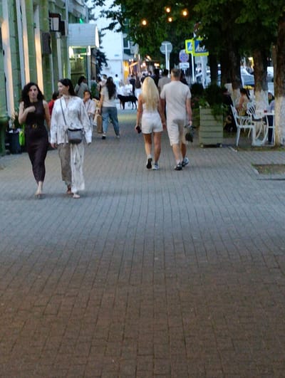 The image depicts a street scene in Maykop, Russia, likely during the evening, given the low light and illuminated string lights visible in the background. A paved walkway, constructed with rectangular gray pavers, dominates the foreground. Several individuals are walking on the pathway. On the left, two women are walking towards the viewer. The woman closer to the viewer wears a dark, possibly purple, dress and the woman to her right wears a light-colored, translucent patterned jacket over light pants and a dark crossbody bag. Further down the path, a couple is walking away from the viewer. The woman has blonde hair and wears white shorts and a white top, while the man wears white shorts and a light-colored t-shirt. Several other people and a black dog are visible in the mid-ground, along with outdoor seating and umbrellas of a cafe on the right side of the image. The background shows buildings and trees, contributing to an urban outdoor setting.