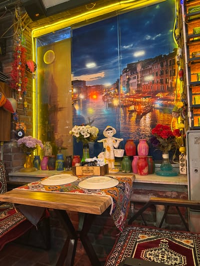 The image depicts an indoor scene, likely a restaurant, with a strong emphasis on colorful and eclectic decor. In the foreground, a wooden table is set with two white plates and a vibrantly patterned runner. To the right of the table, a chair is partially visible, adorned with a patterned rug.
The background is dominated by a large mural of a canal scene in Venice, with buildings lining the water and a twilight sky. Various decorative items are placed on a shelf below the mural, including a small figurine of a child carrying baskets, several colorful vases, and two bouquets of flowers. To the left of the mural, a brick wall is visible, decorated with strings of red tassels and small, colorful ornaments, possibly reminiscent of Nazar amulets. A bright yellow LED light strip runs along the top edge of the wall.
The overall impression is one of a lively and somewhat kitschy ambiance, suggesting a restaurant with a theme that embraces cultural and decorative elements. While the image does not provide direct clues to its exact location within Istanbul, the stylistic choices could suggest a restaurant catering to tourists or a place aiming for a unique, bohemian atmosphere.
