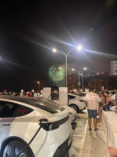 At night in Alicante, Spain, fireworks are erupting in the sky, casting a green and red glow. A crowd of people are gathered at a Tesla charging station. Several white Tesla vehicles are parked, with charging cables plugged in. In the background, buildings and a hillside are visible. Some individuals are looking up at the fireworks, while others are conversing. The scene is illuminated by streetlights and the glow from the charging stations.