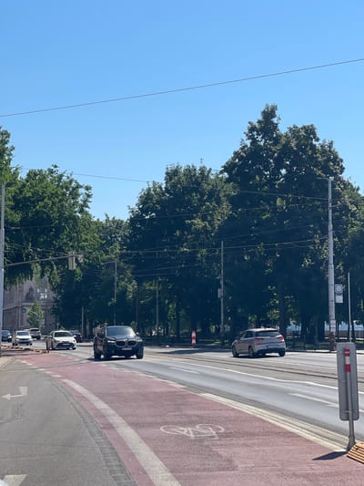 The image depicts a street scene in Bratislava, Slovakia, on a sunny day. The foreground features a red-colored bike lane marked with a white bicycle symbol and directional arrows. Adjacent to the bike lane is a traffic lane with several cars. In the center lane, a dark SUV with license plate "WB 04165" is driving towards the viewer. To its left, a white sedan is visible. Further down the road, a light-colored hatchback is moving away from the viewer. The background is dominated by a row of lush green trees, and a clear blue sky. Utility poles and wires are strung across the scene, and a bus stop sign is visible on the right side. The overall impression is of a busy urban environment with dedicated infrastructure for cyclists.