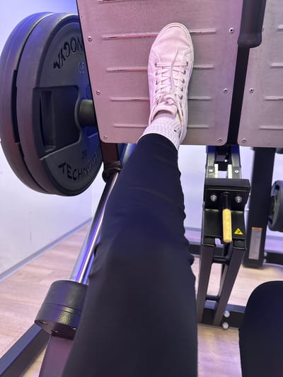 In the foreground, a person's legs are visible, clad in black leggings, with a white sneaker and white socks on their right foot. The foot is positioned on the platform of a weight machine, likely a leg press. To the left, a large, circular weight plate with "TECHNOGYM" and "20" stamped on it is partially visible, indicating a weight of 20 kilograms. The machine itself is made of metal and has a black and silver color scheme. The background shows a gym environment with a light-colored floor and a wall illuminated with purple and blue lighting. There is no identifiable information about the specific location in Helsinki, Finland, as the image contains no discernible landmarks or context clues to pinpoint it within the city. The scene depicts a solitary individual engaged in a weightlifting exercise.