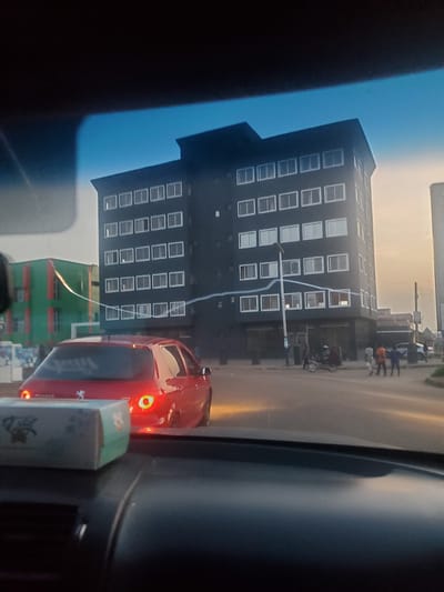 The image captures a view from inside a vehicle in Sardauna, Nigeria, showing a street scene at dusk. A dark, modern, multi-story building with numerous rectangular windows dominates the background. To the left, a green building with orange vertical accents is visible. In the foreground, the rear of a red Peugeot hatchback is prominent, with its taillights illuminated. A tissue box rests on the dashboard in the lower left corner. Several figures are seen walking and standing on the street near the dark building, and a motorcycle is parked in the vicinity. The sky is a soft, hazy gradient of orange and blue, indicating either sunset or sunrise.