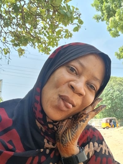 This outdoor portrait features an individual, likely a woman, looking directly at the camera with her head tilted to the left. She is wearing a black head covering, possibly a hijab, patterned with bold red and earthy-toned abstract shapes. Her complexion is warm, and she has a small nose stud on her right nostril. Her expression appears calm, with her tongue slightly visible.
Her right hand, positioned near her chin, displays intricate, dark brown mehndi or henna designs across the back of her hand and fingers, featuring geometric and possibly floral patterns. A dark wristwatch is visible on her left wrist.
The background is bathed in bright daylight under a clear blue sky, indicating fair weather. Lush green trees and foliage are visible in the upper right and lower left parts of the frame. Faint utility lines can be seen strung across the sky. In the blurred lower left background, a yellow auto-rickshaw (often referred to as a tuk-tuk) is partially visible, suggesting an urban or semi-urban street setting. The image itself is rotated, giving the composition an angled perspective. The presence of the auto-rickshaw and the overall environment align with a location like Shaba, Nigeria. No discernible text is visible in the image.