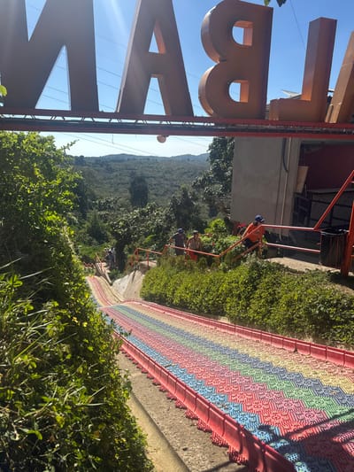 A vibrant, rainbow-colored patterned wall runs alongside lush green vegetation, with workers ascending a staircase alongside it. In the distance, a verdant mountain landscape fills the horizon. A partially visible sign with large orange letters is in the foreground, framing the scene. The image is oriented at an unusual angle.