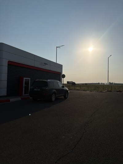 The image, when viewed in correct landscape orientation, captures a bright, sunny daytime scene featuring a modern commercial building, a dark-colored SUV, and a vast open field adjacent to a paved area.
In the upper left portion of the image, the sun shines brightly against a clear, light blue sky, casting a strong glare and long shadows, indicating either morning or late afternoon. Several tall streetlights are silhouetted against the sky, with one prominent pole extending upwards from the middle of the frame.
To the left, a contemporary building with gray paneling and a horizontal red stripe stands prominently. The building features vertically ridged sections and a window with a red frame. A security camera is mounted on the building's exterior. Parked in front of the building, with its rear facing the viewer, is a dark SUV or crossover, distinguished by a spare tire mounted on its tailgate. The car's license plate is clearly visible and reads "A 9600 KM", indicating it is a Bulgarian vehicle (Sofia region code 'A').
The foreground and midground on the right side of the image are dominated by an asphalt surface, likely a parking lot or a section of a road. Red and white curb markings delineate the edge of the paved area near the building. A distinct crack runs diagonally across the asphalt in the lower right. Beyond the paved area, a long stretch of green and golden field, possibly farmland, extends into the distance, bordered by a fence. Faint structures, possibly a distant bridge or other buildings, are visible on the horizon under the bright sky.
The scene depicts a static moment at what appears to be a roadside stop or service area in a semi-rural setting near Vodenichane, Bulgaria. There are no people visible, and the overall impression is one of calm and stillness under clear, sunny weather.