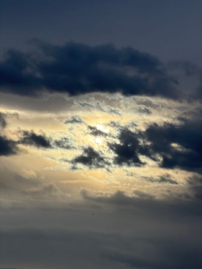 The image shows a dramatic sky filled with dark, heavy clouds. Sunlight is breaking through the clouds in the center of the frame, creating a bright, illuminated band that highlights the texture and movement of the clouds. The overall mood is somber, with a sense of impending weather or the aftermath of a storm. The scene is entirely focused on the sky, with no visible ground, buildings, or any indication of the city of Leudelange, Luxembourg.