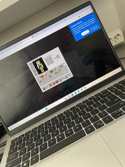 A laptop is open, displaying a Minecraft game. The crafting menu is visible with materials placed for a character skin. A Chrome browser with multiple tabs is open in the background, with some tabs labeled in a Cyrillic language. A keyboard sits to the right of the screen. A two-plug electrical outlet is nearby.