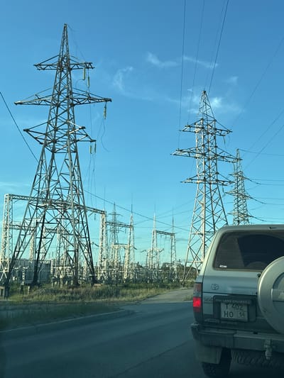 The image depicts a landscape dominated by a high-voltage electrical substation under a clear blue sky with scattered white clouds. Several large steel lattice towers, supporting power lines, are prominent in the foreground and midground. These towers are interconnected by numerous wires, creating a complex network. In the background, a dense arrangement of similar structures and equipment signifies a substantial industrial power infrastructure.

To the right of the substation, the rear of a silver Toyota Land Cruiser is visible, with its spare tire mounted on the tailgate. The vehicle's license plate is clearly legible, displaying "T 402 HO 14" and the text "TOYOTA LAND CRUISER VX LIMITED". The presence of the vehicle and the road suggest the photo was taken from a car driving past or near the substation.

The scene is brightly lit, indicating it is daytime, likely late afternoon or early morning given the long shadows and the quality of light. The sky is a vibrant blue, and the weather appears clear and fair. The setting is industrial, characterized by the electrical infrastructure, and the location is identified as Yakutsk, Russia. No people are visible in the image, and the overall mood is one of industrial efficiency and vastness. The image is oriented vertically, with the sky at the top and the ground at the bottom.