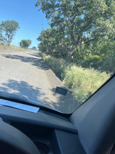 The image shows a view from inside a vehicle looking out a windshield at a dusty, unpaved road winding through a landscape with trees and vegetation. The road appears to be a rural track. In the foreground, part of the vehicle's dashboard is visible, with a piece of white paper lying on it. On the right side of the road, a dark rectangular object with a circular logo is resting on the gravel shoulder. The sky is clear and blue. There is no indication of the city of Poroy, Bulgaria, in the image.