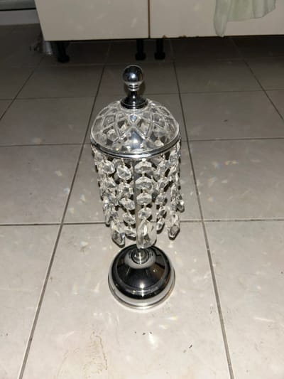 An indoor scene in Marousi, Greece, depicts a stationary decorative object composed of chrome and crystal. The object stands centered on a light-colored tiled floor, featuring square ceramic tiles with dark grout lines.
The decorative object consists of a polished chrome circular base, supporting a central chrome stem. Attached to the stem is a faceted clear crystal dome, from which multiple strands of clear, faceted crystal pendants cascade downwards. A small, spherical chrome finial caps the crystal dome. Reflections off the chrome surfaces and refractions from the crystals are visible on the surrounding tiles.
In the background, two light-colored cabinets or furniture pieces with dark, cylindrical support feet are visible. A light-colored fabric or textile hangs partially into view on the right side, behind the decorative object and against the cabinetry. The scene is devoid of people or dynamic actions.